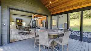 A wooden deck with a round table and wicker chairs, leading to a kitchen area with a staircase. - Discover the perfect escape in our brand-new, secluded cabin in nature's embrace (Blue River)