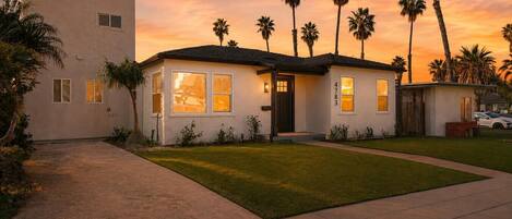 A single-story house with a front yard, palm trees, and a sunset sky.