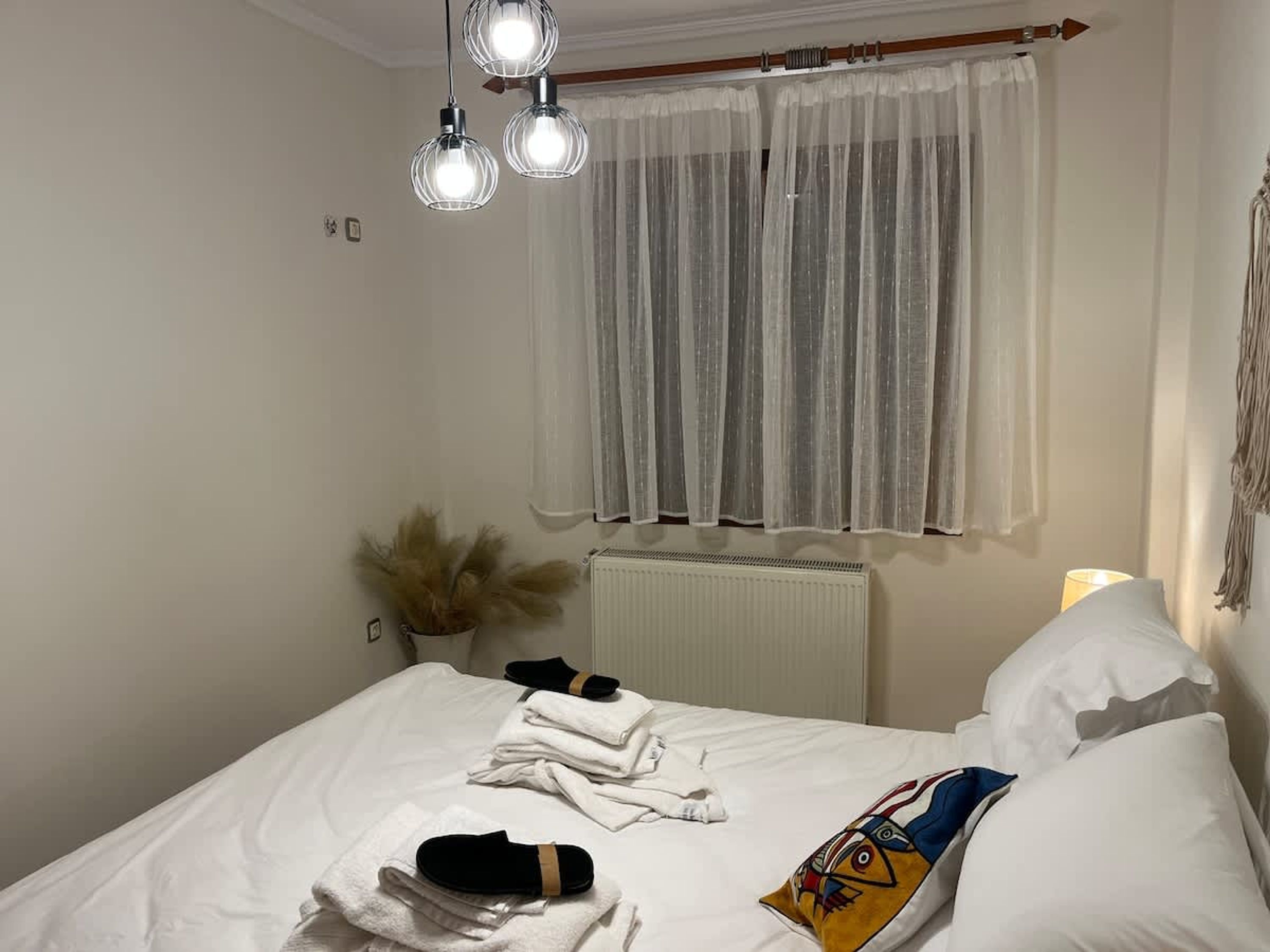 A neatly made bed with towels and slippers, a radiator, a window with sheer curtains, and a hanging plant.