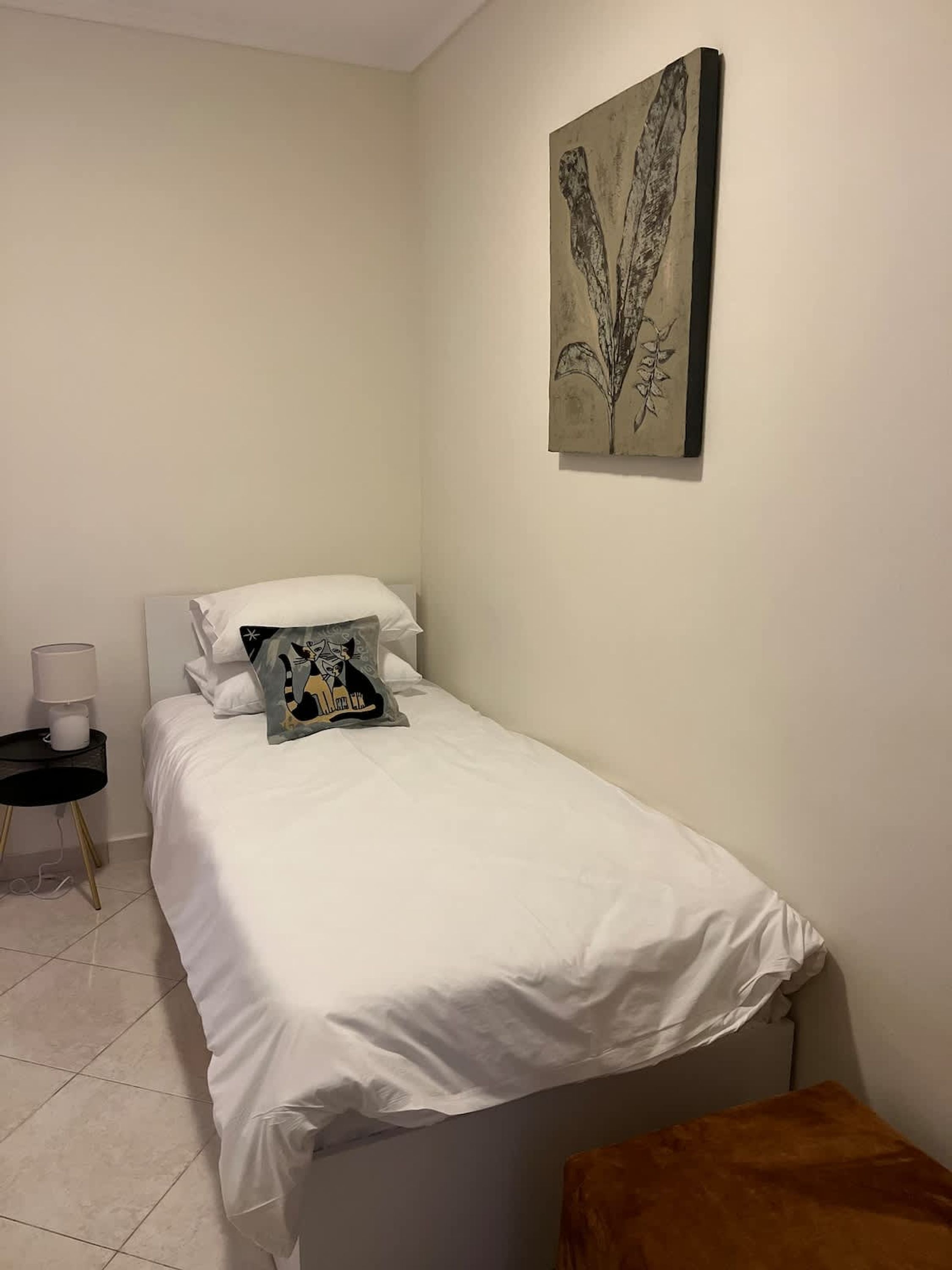 Une petite chambre à coucher, bien rangée, avec un lit simple, une table de chevet avec une lampe et un petit tabouret.