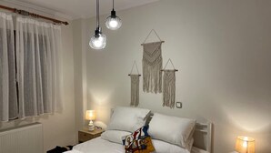 Een net opgemaakt bed met witte lakens, een kleurrijk kussen en handdoeken ernaast. Twee bedlampen en macramé wanddecoraties zijn zichtbaar.