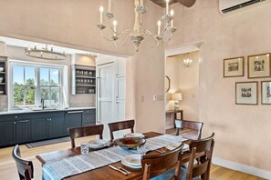 Un coin repas avec une table en bois dressée pour quatre personnes, entourée de chaises. Un lustre est suspendu au-dessus de la table, et une fenêtre offre une vue sur la verdure à l’extérieur.