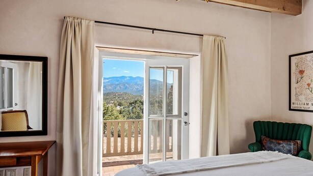A bedroom with a large bed, a green chair, a wooden desk, and a balcony with a view of mountains. - Santa Fe Santuario- Perfect Views, Kiva Fireplace (Santa Fe)
