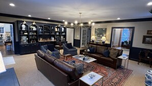 Een ruime woonkamer met een donkerblauwe bank, een bijzettafeltje en een kamerkleed met een patroon. Er is een boekenkast vol decoratieve objecten en een kroonluchter hangt aan het plafond.