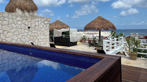 A pool area with a stone wall, a thatched umbrella, and a view of the ocean. - Walk to Waterfront! Luxury King Suite w/ Rooftop Pool & Spa (Cozumel)