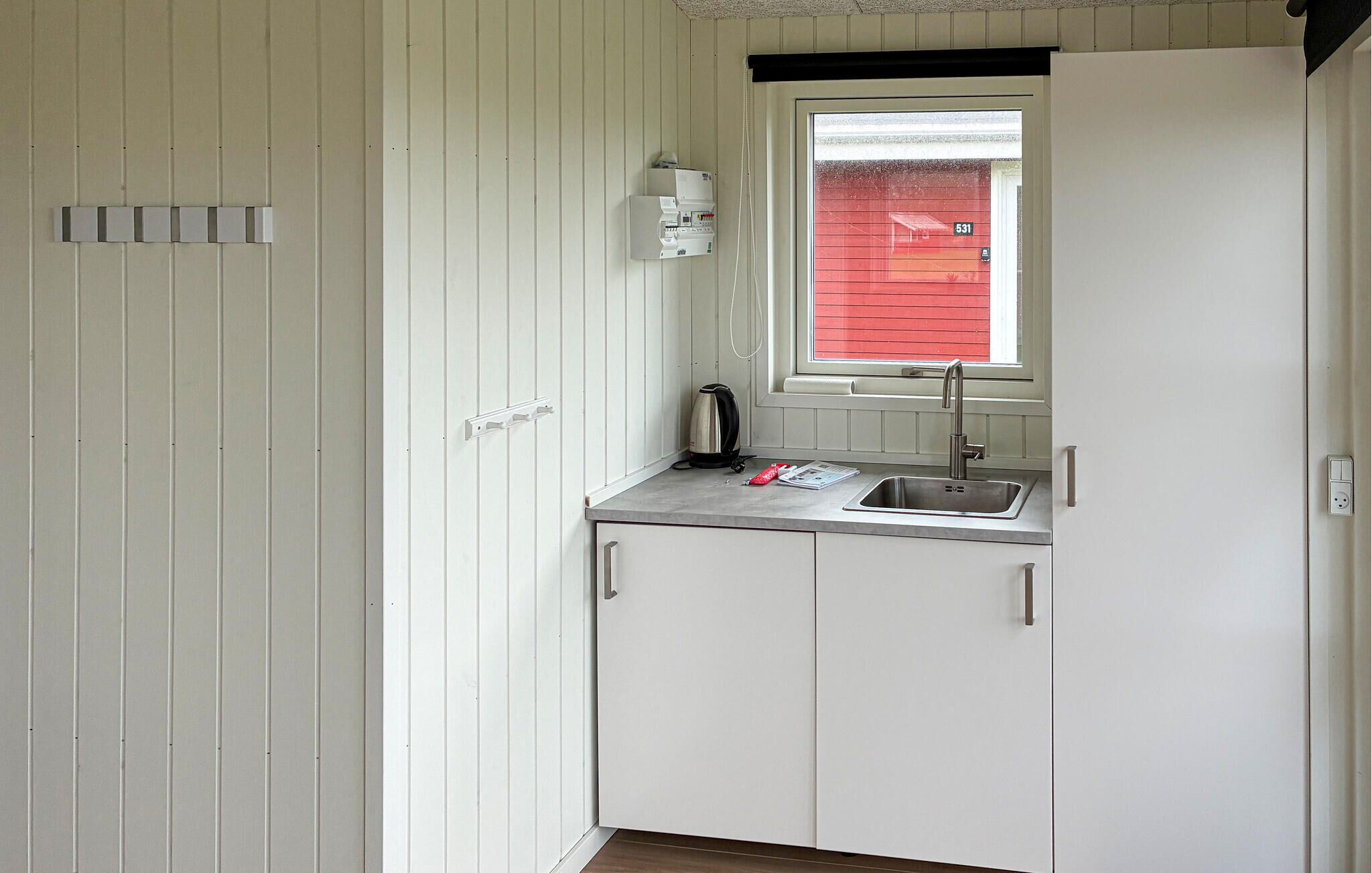 A compact kitchen with white cabinets, a window with a red exterior, a kettle, and a sink.