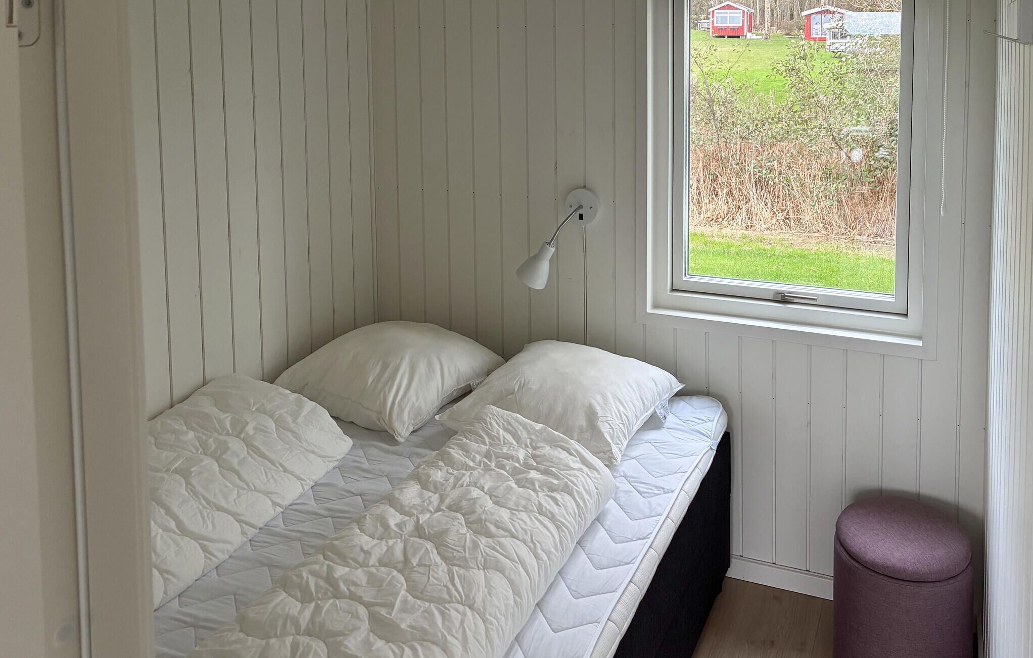 Een eenpersoonsbed met een witte quilt, een bedlampje en een raam met uitzicht op een rode woning en groen buiten.