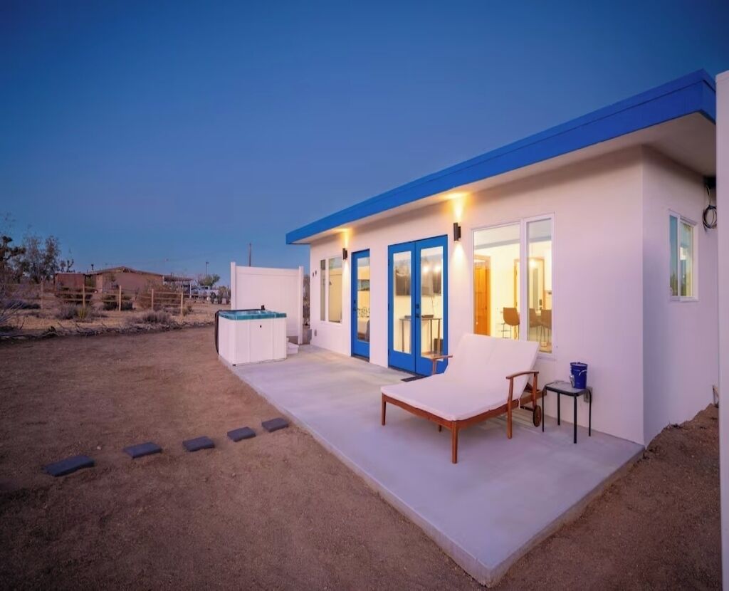 A modern building with a blue roof and white walls, featuring a covered patio with a lounge chair and a small table.