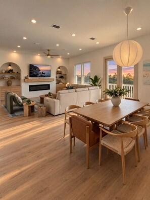 Una sala de estar moderna con una mesa de comedor de madera, asientos cómodos, un televisor de pantalla plana y un ventanal con vistas al atardecer.