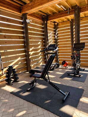 Ein überdachter Outdoor-Fitnessbereich mit Trainingsgeräten, darunter eine Bank, Kurzhanteln und ein Ergometer.