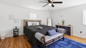Ein Schlafzimmer mit Bett, Deckenventilator, Nachttisch, Sitzbank, blauem Teppich und einer kleinen Pflanze auf einem Schrank.