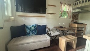 A cozy living area with a sofa, a TV mounted on the wall, a small wooden table with two wine glasses, and a window with curtains. - Private Tiny Home Retreat Easy Fort Worth Access (Azle)