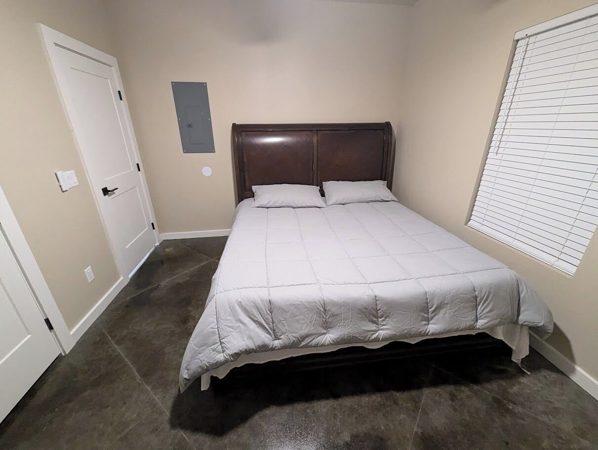 A bedroom with a bed, a door, a light switch, and a window with blinds.