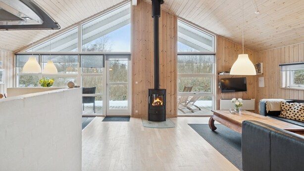 A modern interior with a wood-burning stove, large windows, and a cozy seating area. - Holiday home with spa and sauna, close to nature (Knebel)
