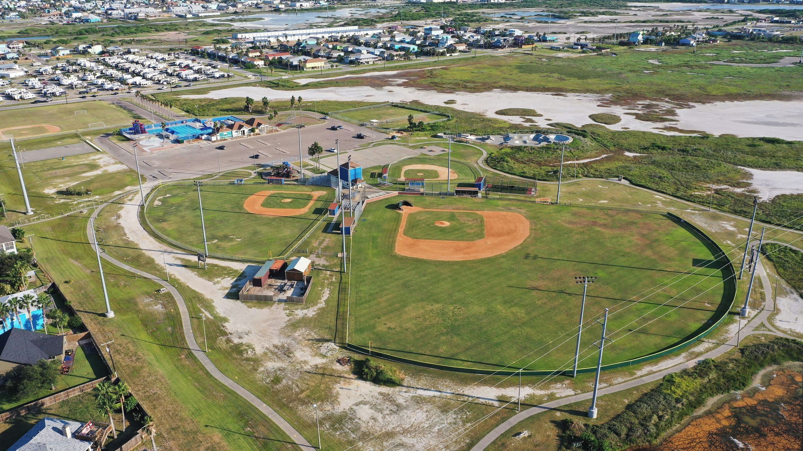 An aerial view of a baseball field with multiple infield diamonds, a dugout, and a parking area.
