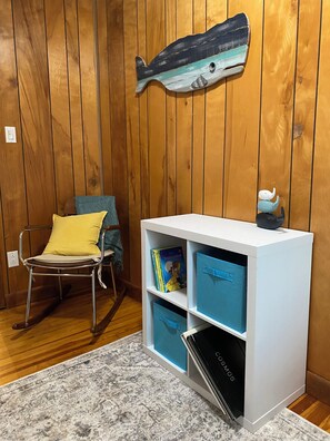 A room with wooden paneling, a rocking chair with a yellow cushion, a white bookshelf with blue storage bins, and a fish-shaped decoration on the wall.