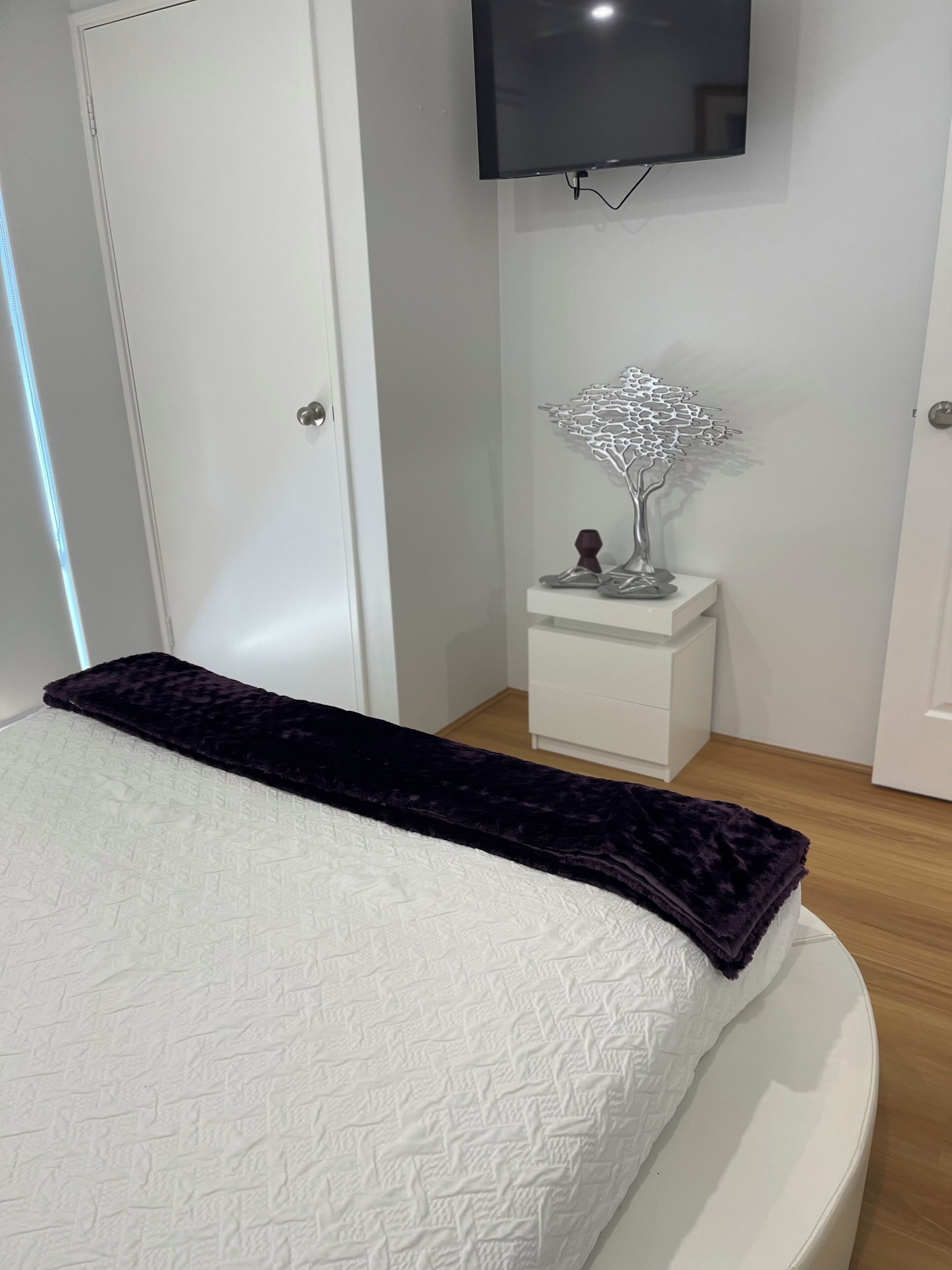 Una camera da letto con un letto, un comodino bianco, un albero decorativo in metallo e una TV a schermo piatto montata a parete.