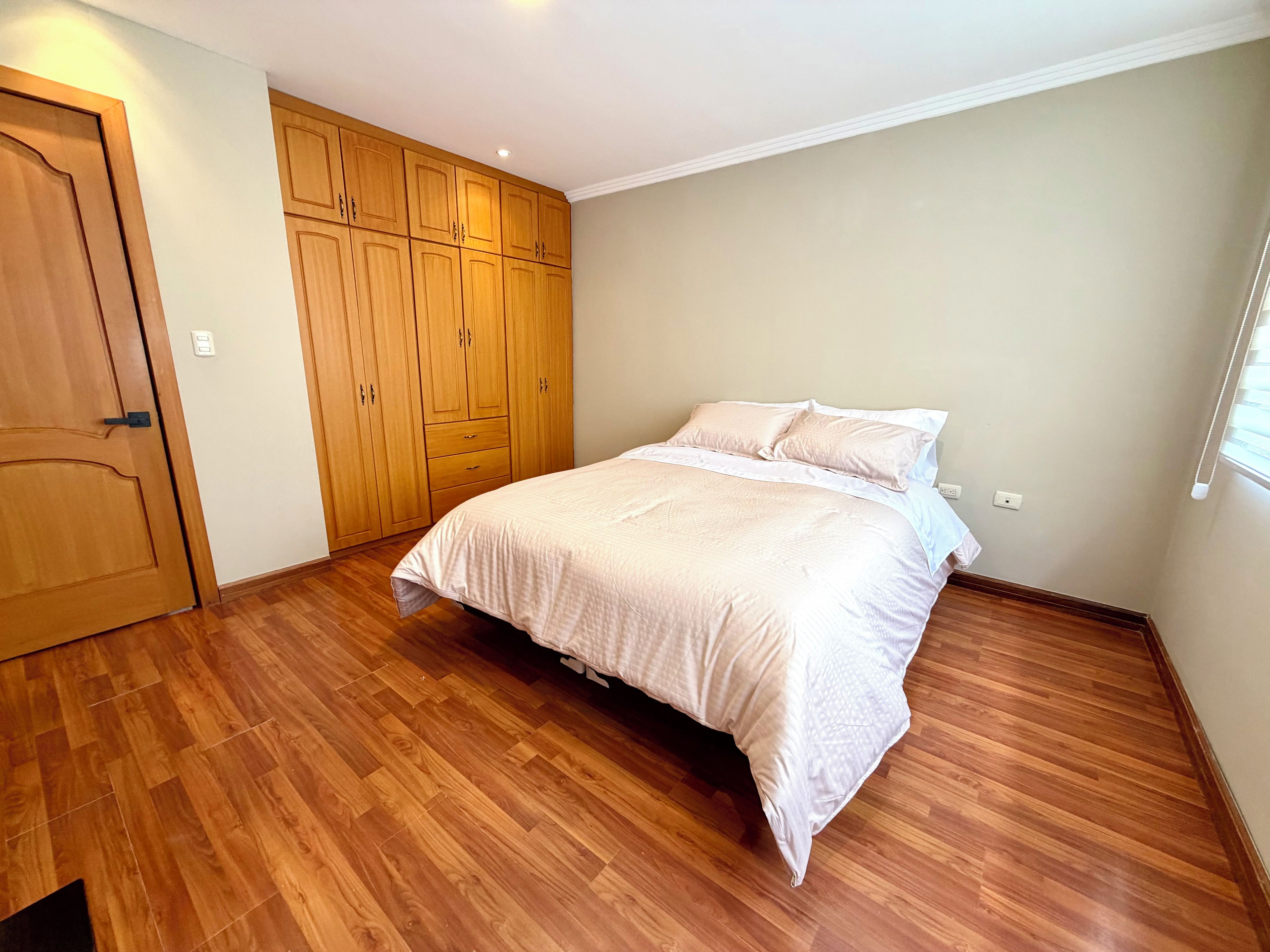 A bedroom with a wooden wardrobe, a bed with white bedding, and a wooden floor.