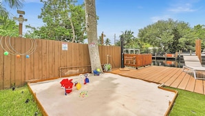 Un jardin avec une terrasse en bois, un espace de jeux sablonneux et une clôture en bois.
