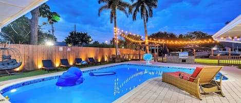 A swimming pool with lounge chairs and a barbecue area, surrounded by palm trees and string lights.