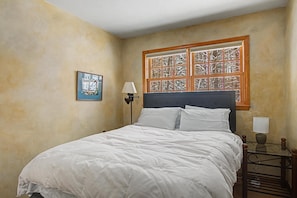 Une chambre à coucher avec un lit, une table de chevet avec une lampe, une fenêtre ornée d’une grille décorative et un tableau encadré accroché au mur.