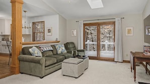 Un salón con un sofá verde, un reposapiés y una mesa de centro de madera. Se ve una ventana grande con vistas a árboles cubiertos de nieve.
