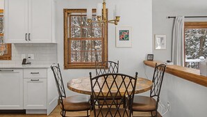 Una cocina con armarios blancos, una mesa redonda de madera con cuatro sillas, una araña y una ventana con vistas a árboles cubiertos de nieve.