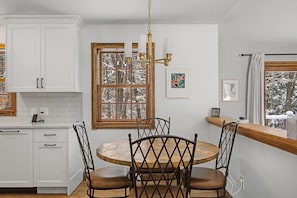 Une cuisine avec des meubles blancs, une table ronde en bois entourée de quatre chaises, un lustre et une fenêtre donnant sur des arbres couverts de neige.