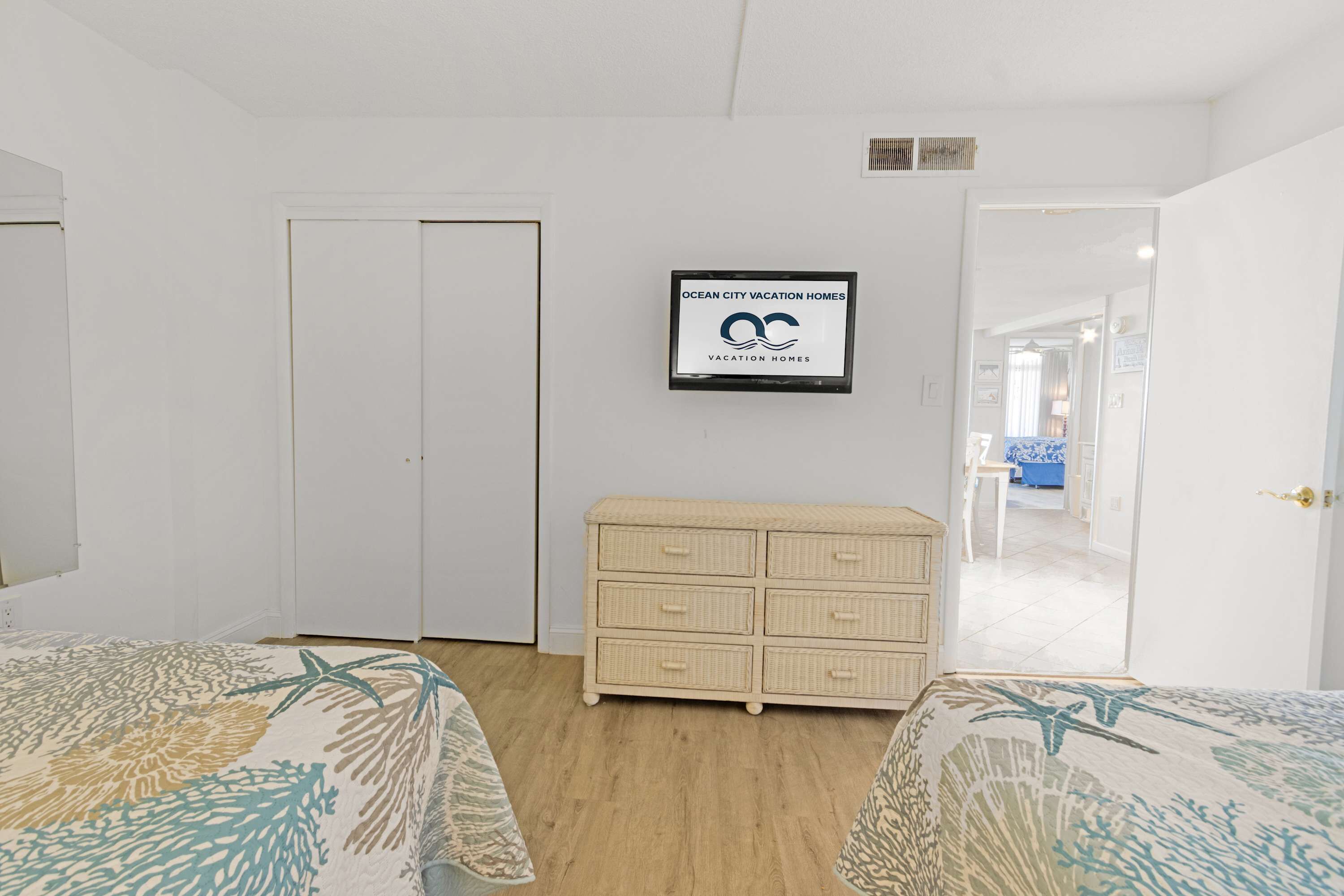 A bedroom with two beds, a white wardrobe, a framed sign, and a view into another room.
