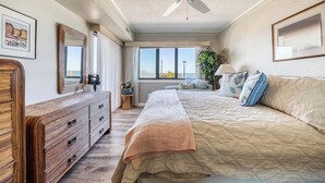A bedroom with a bed, a dresser, a ceiling fan, a large window, and a view of the ocean. - Plaza 109 (Ocean City)