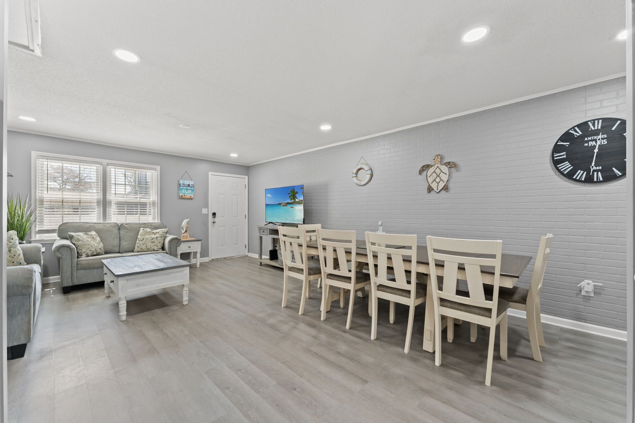 Una sala de estar con un sofá, una mesa de centro y una mesa de comedor con sillas. Hay un reloj en la pared y un cuadro sobre la mesa de comedor.