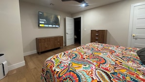 Un dormitorio con una cama grande, ventilador de techo, televisor montado en la pared, cómoda de madera y armario.