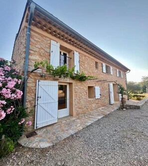 A stone building with a gravel driveway, white shutters, and a small garden with pink flowers. - LES CLAIRIANS - LE CANNET DES MAURES (LE CANNET DES MAURES)