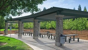 Un'area picnic riparata con colonne in pietra e panche, circondata da alberi e un vigneto.