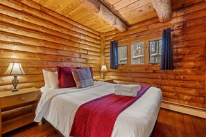 A log cabin bedroom with a wooden bed, two bedside tables with lamps, and a window with curtains. - Stratton Log Lodge: Hot Tub + Privacy | Sleeps 13 (Jamaica)