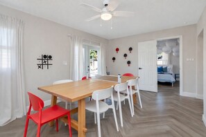 A room with a wooden dining table, white chairs, a ceiling fan, and a bed with blue bedding. - Poolside Playhouse - Pool Home - 5 Min. To Beach (Seminole)