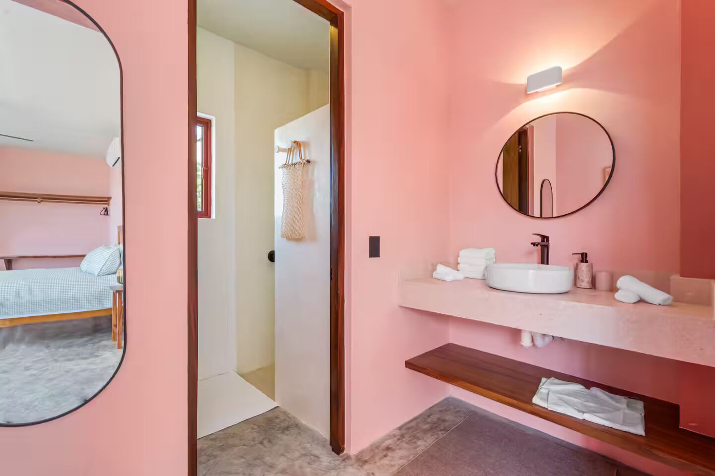 A modern bathroom with a pink wall, a round mirror, a white sink, and a wooden shelf.