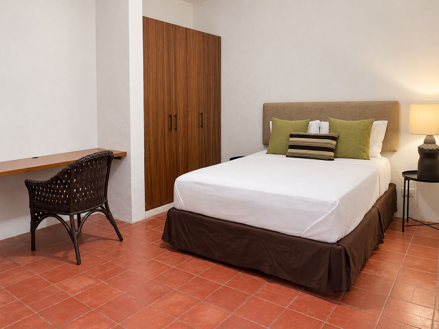 A hotel room with a bed, a desk, a chair, a wardrobe, and a nightstand with a lamp.