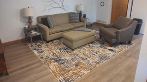 Uma sala de estar com sofá, um apoio para os pés, uma poltrona, mesa de centro, mesa lateral, abajur, obra de arte na parede e uma porta.