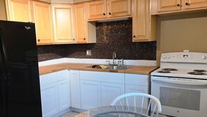 Une cuisine avec des meubles en bois, un réfrigérateur noir, une cuisinière blanche, une table à manger en verre et un ventilateur de plafond.