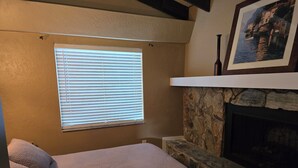 Une chambre à coucher avec un lit, une fenêtre avec des stores, une cheminée en pierre et un tableau encadré au-dessus de la cheminée.