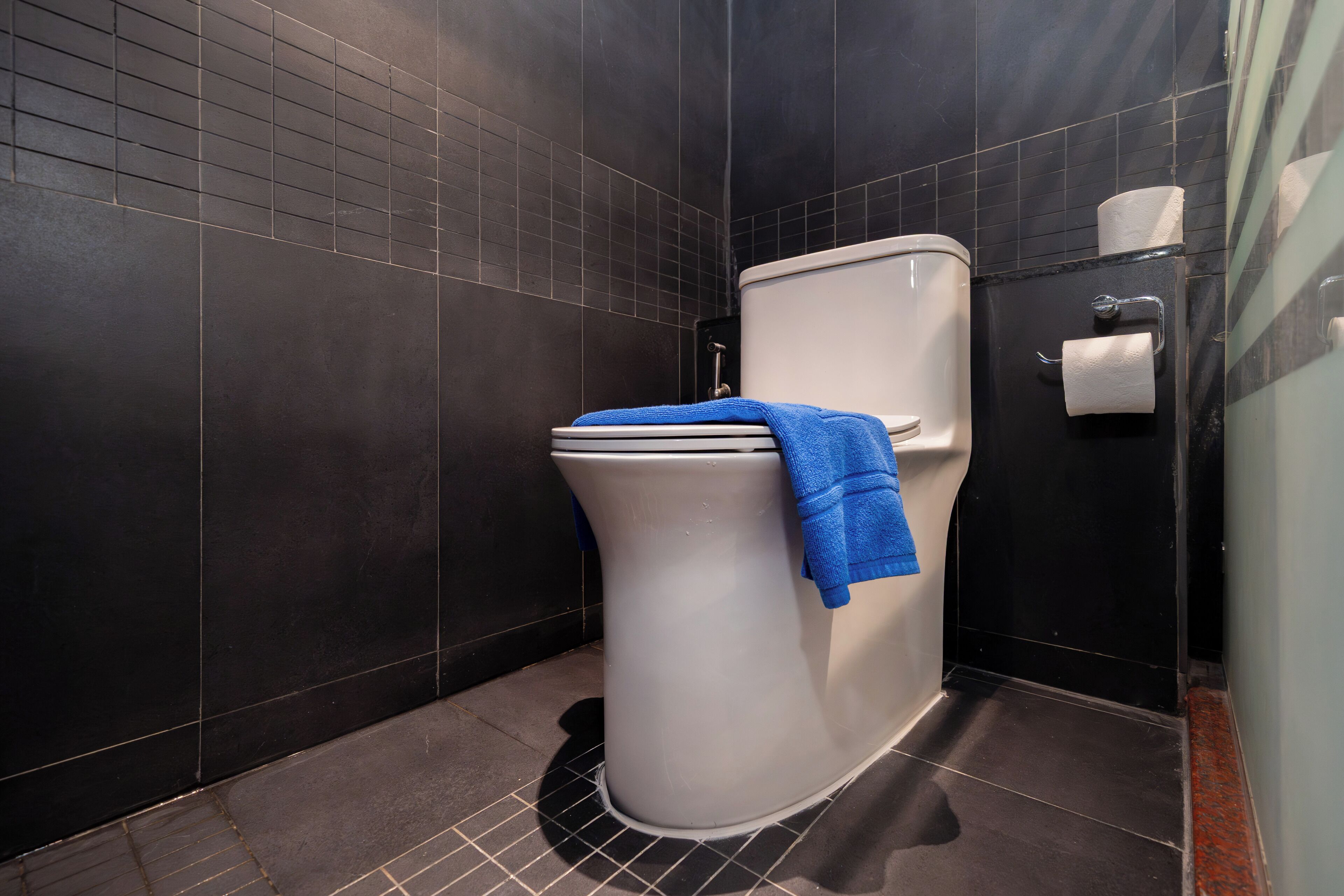 A modern bathroom with a white toilet, dark tiled walls, and a blue towel on the seat.
