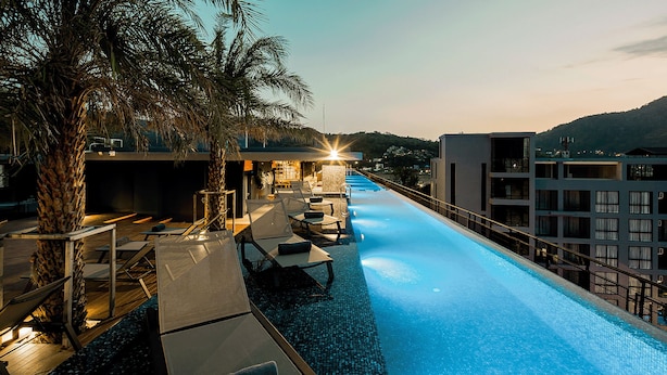 A swimming pool with lounge chairs and a palm tree, surrounded by buildings. - Chic N Cozy Citygate 1Br Suite (Kamala)