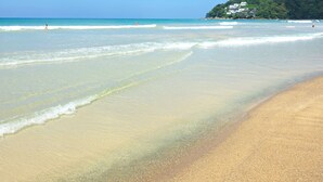 A beach with clear water, a sandy shore, and a hill with buildings in the background. - Chic N Cozy Citygate 1Br Suite (Kamala)