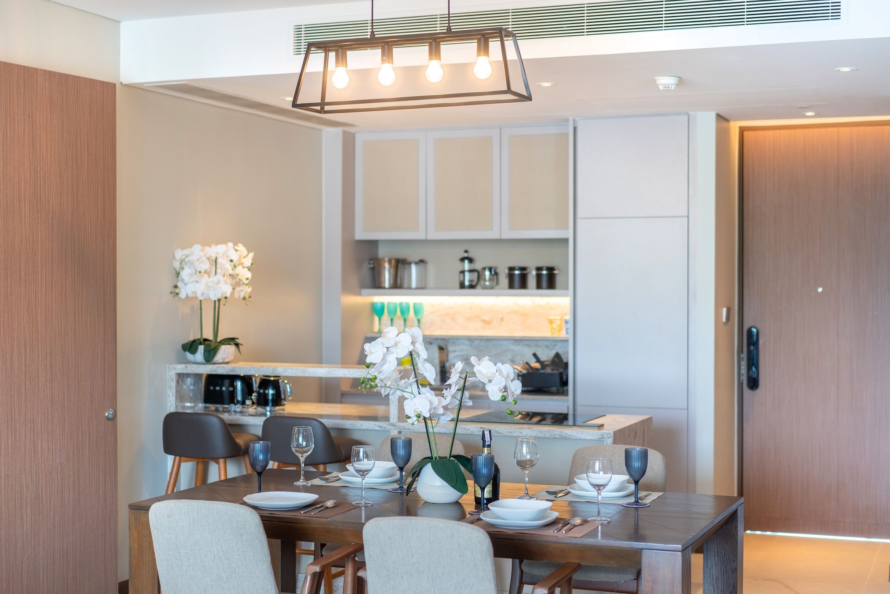 A modern dining area with a wooden table set for four, a vase of white orchids, and a kitchen in the background with white cabinets and a countertop.