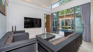 A modern living room with a flat-screen TV, wicker furniture, and large windows offering a view of a swimming pool and greenery. - Mahogany 4Br Pool Villa In Bangtao (Choeng Thale)