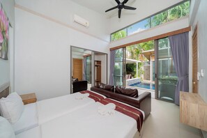 A hotel room with a bed, a sofa, a small table, a ceiling fan, and a view of a swimming pool. - Mahogany 4Br Pool Villa In Bangtao (Choeng Thale)