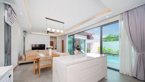 A modern living room with a sofa, dining table, and a TV. There is a large window with a view of the outdoors. - Nicky Elegant 3Br Pool Villa Bangtao (Choeng Thale)