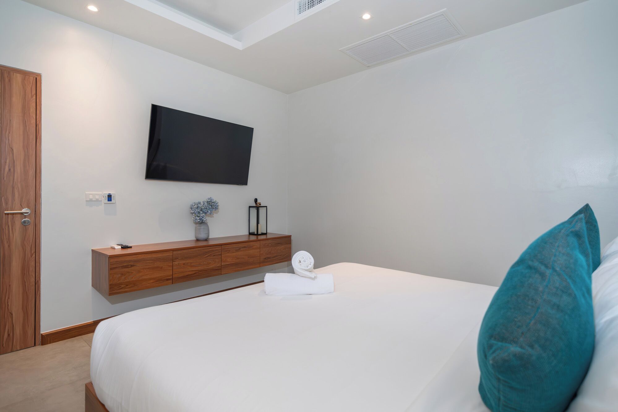 A bedroom with a bed, a TV mounted on the wall, a wooden bedside table, and a wall-mounted air vent.