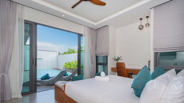 A modern hotel room with a large bed, a ceiling fan, a desk with a chair, and a view of a swimming pool and greenery outside. - Nicky 3Br Private Pool Villa Bangtao (Choeng Thale)